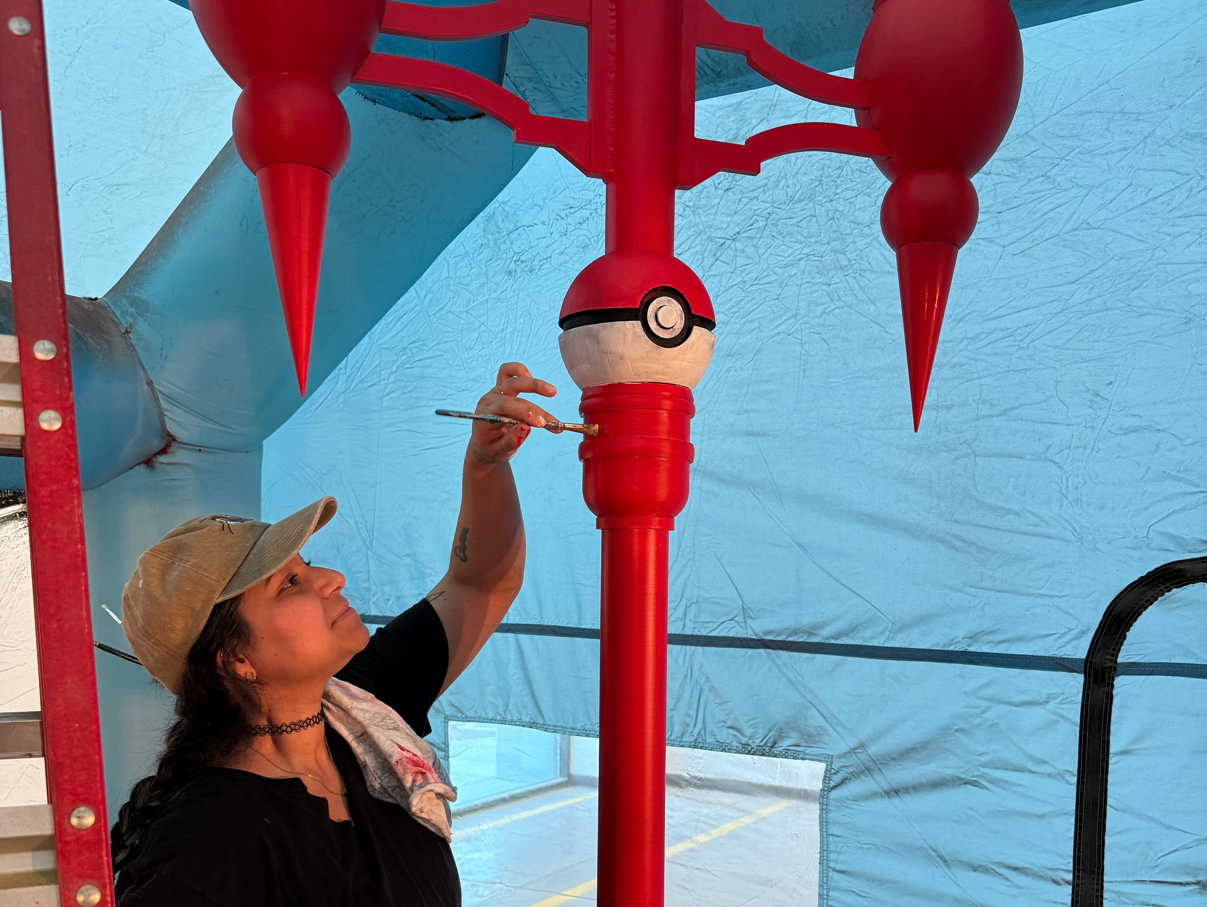 Andy Aby painting a red and white Pokéball-themed decorative lamp-post under a light blue tent. The pole features three red fin-like tops and a central Pokéball design.