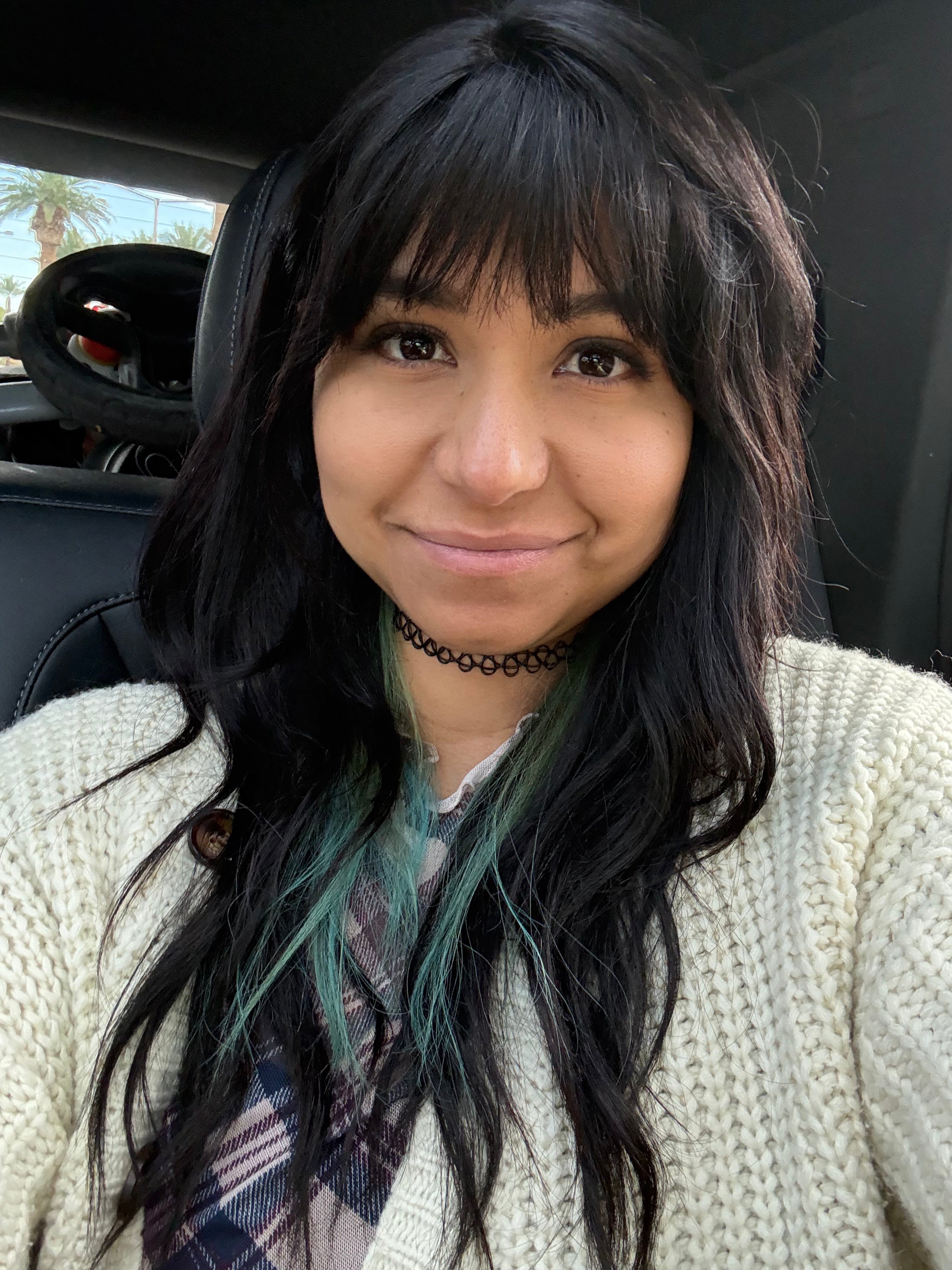 Andy Aby, Female-presenting with long black hair featuring a green-tinted underlayer, wearing a cream-colored chunky knit cardigan and a plaid shirt, sitting inside a car.