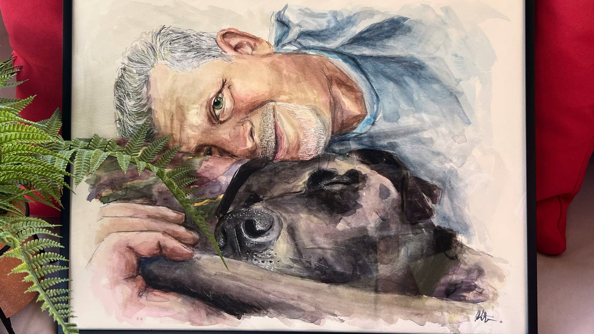 A framed custom watercolor portrait of an older man and his dog, displayed on a sofa with red cushions and a green plant in the foreground.
