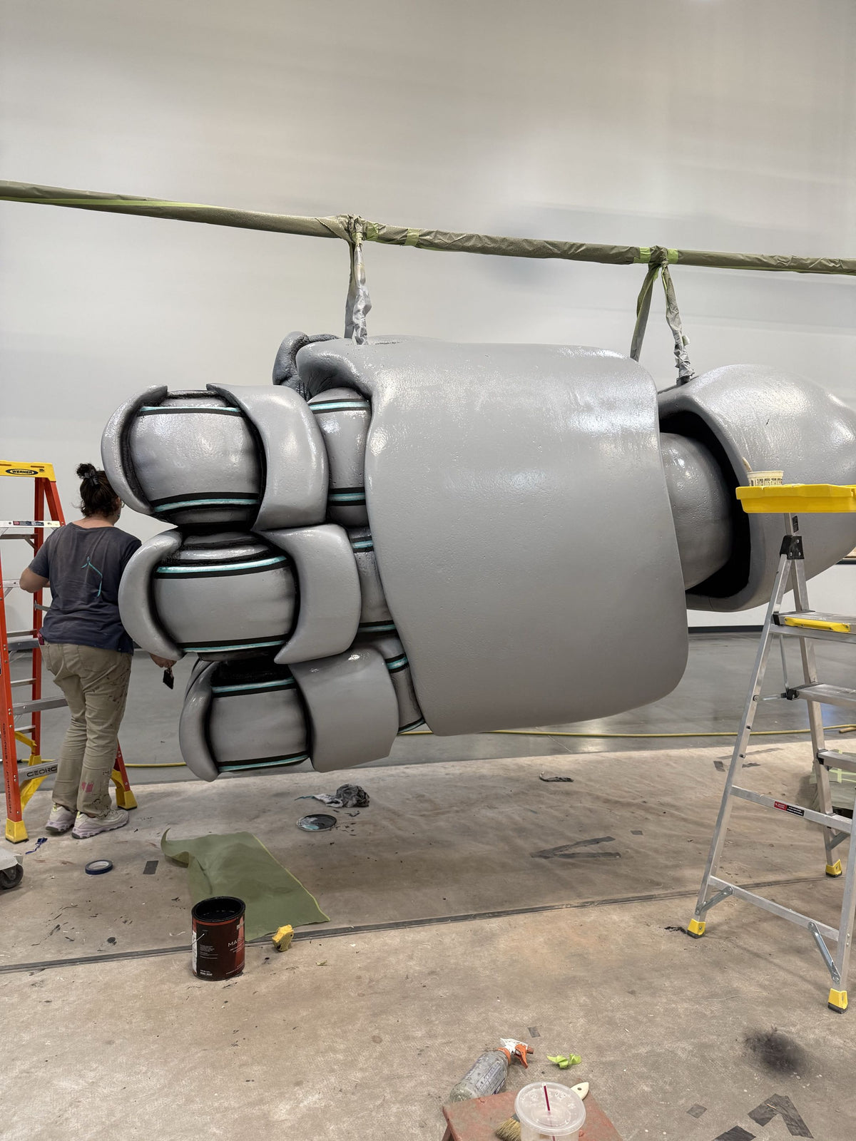 A large, custom-built sci-fi style mechanical claw or fist prop, painted in a matte gray finish, suspended from a crane in a workshop. A person is visible on the left working on another structure, and ladders are present on both sides.