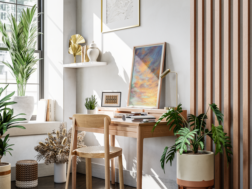 A styled home office corner featuring a light wood desk and matching chair, decorated with a colorful landscape painting, a brass desk lamp, books, and several green potted plants. The background includes a white wall with a floating shelf, a slatted wood screen, and a large window.