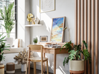 A styled interior corner with a wooden desk and chair, potted plants, a shelf with decorative objects, and a framed abstract painting leaning against a light wall.
