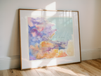 A framed watercolor art print leaning against a white wall on a light wood floor, featuring soft pastel clouds in purple, pink, blue, and orange tones.