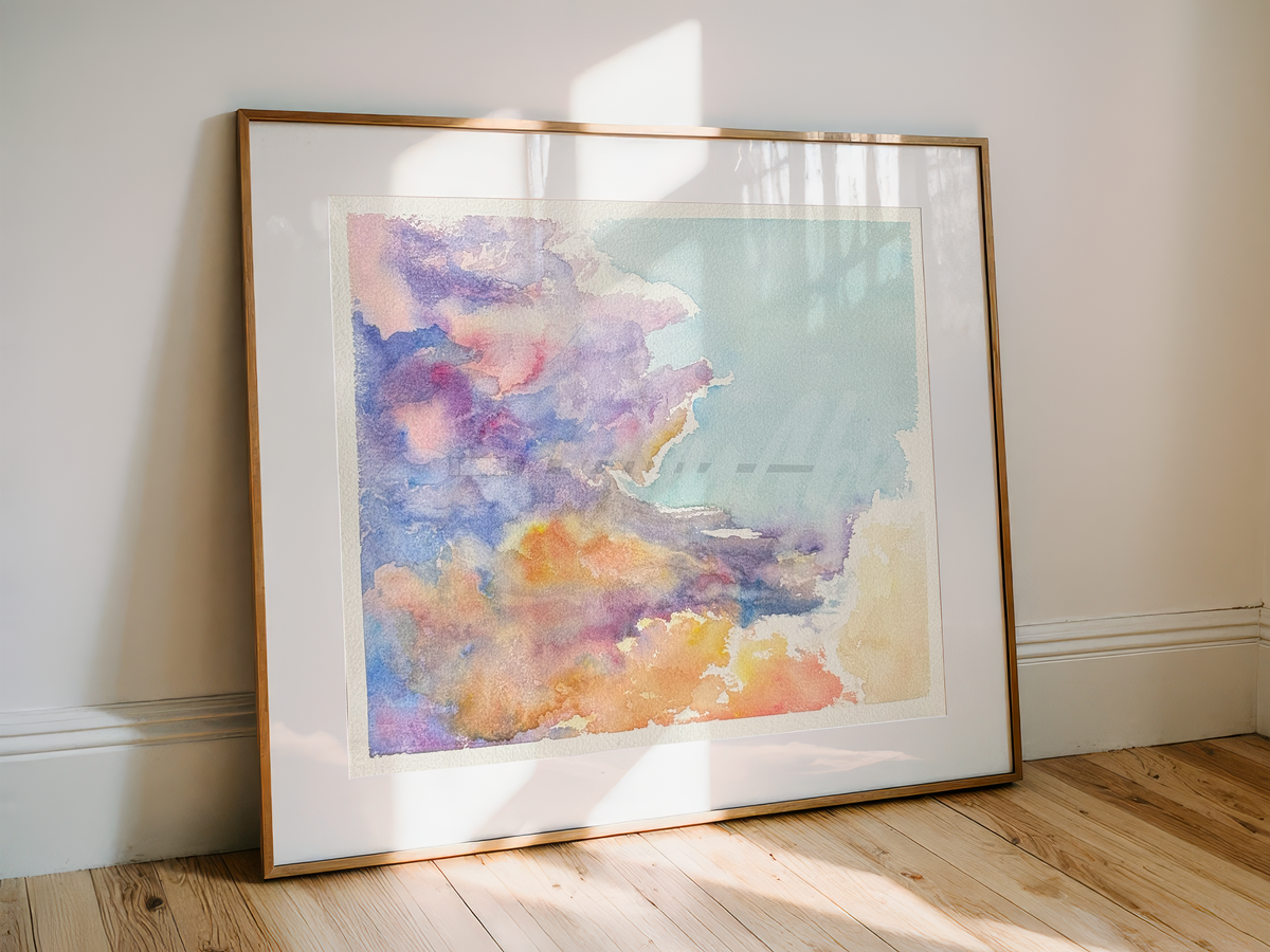 A framed watercolor art print leaning against a white wall on a light wood floor, featuring soft pastel clouds in purple, pink, blue, and orange tones.