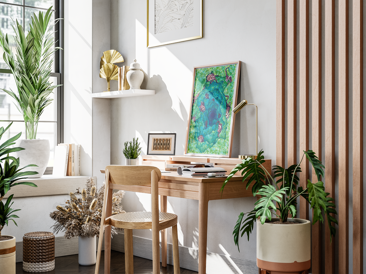 A beautifully styled home office corner featuring a light wood desk and matching chair, decorated with a colorful framed art print, a brass desk lamp, books, and potted green plants in a bright, sunlit room.
