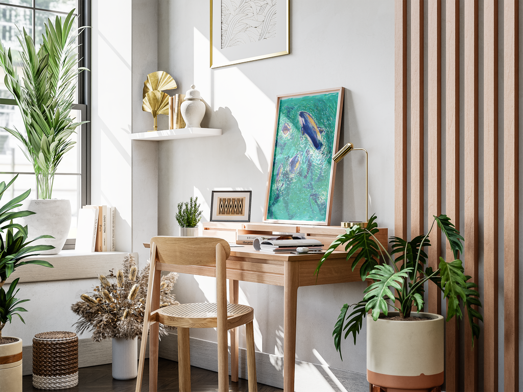 A styled interior corner with a light wood desk and matching chair, decorated with framed art, a gold fan-shaped wall decor piece, books, dried grasses, and several green houseplants in a bright room with natural sunlight.