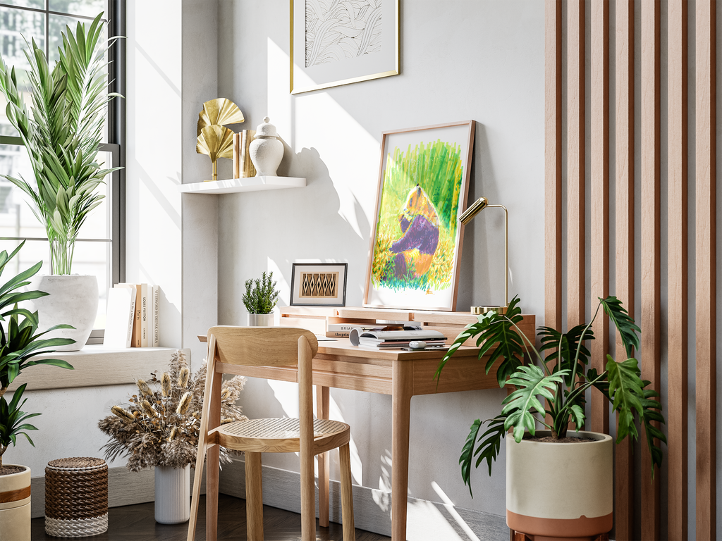 A styled home office corner featuring a light wood desk and matching chair, decorated with a colorful framed art print, a gold desk lamp, and surrounded by lush green potted plants in a bright, sunlit room.