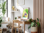 A styled home office corner featuring a light wood desk and matching chair, decorated with a colorful framed art print, a gold desk lamp, and surrounded by lush green potted plants in a bright, sunlit room.