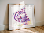 A framed watercolor art print of a tiger's face, leaning against a white wall on a light wood floor. The print features a colorful watercolor tiger portrait with purple, pink, blue, and yellow tones, set in a white mat and a thin gold frame.